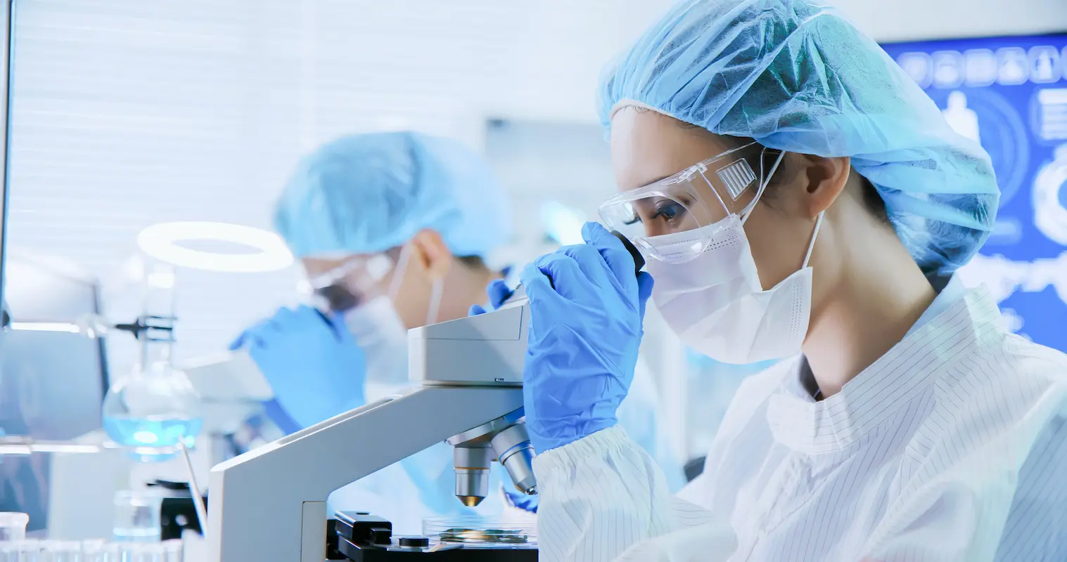 Two scientists in protective clothing work in a laboratory, one using a microscope, while the other appears focused on a different task.
