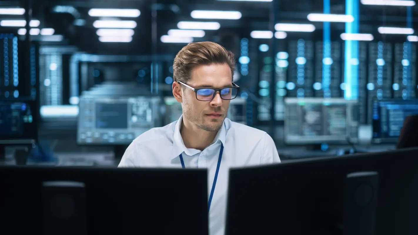 A person is working on computers in a modern office with multiple screens and servers in the background.