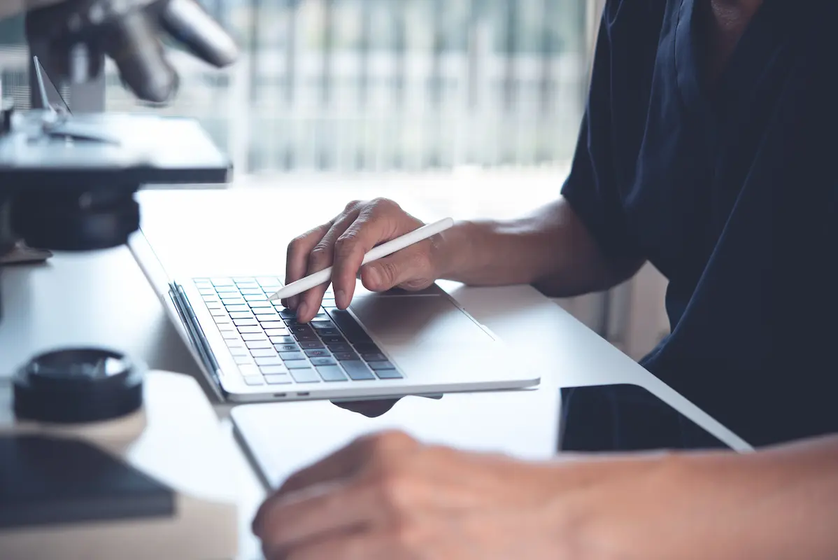 A person uses a stylus on a laptop trackpad. A microscope is positioned on the desk to the left of the laptop.