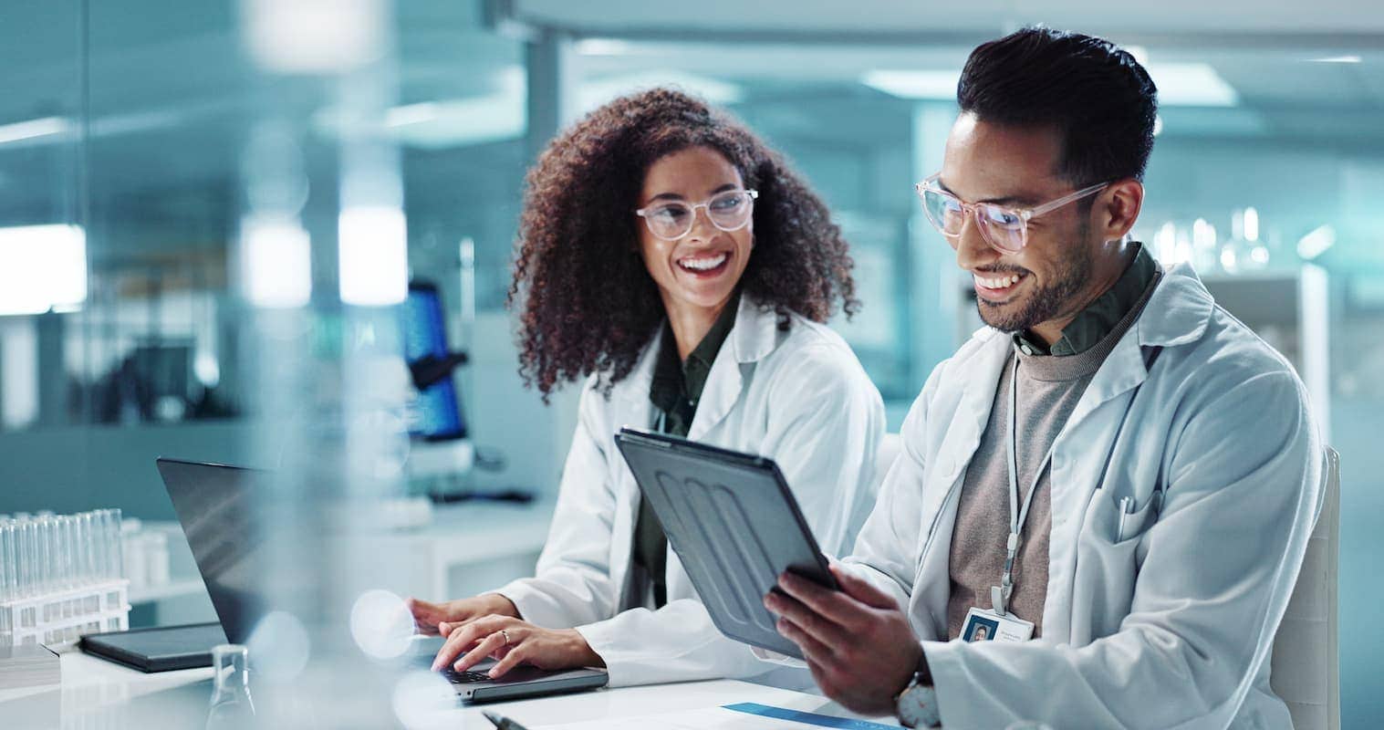 Two scientists wearing lab coats and safety goggles work together in a laboratory; one using a laptop and the other holding a tablet.