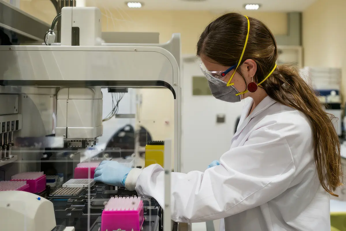 A technician in a lab coat, safety goggles, and a mask operates laboratory equipment with trays of samples.