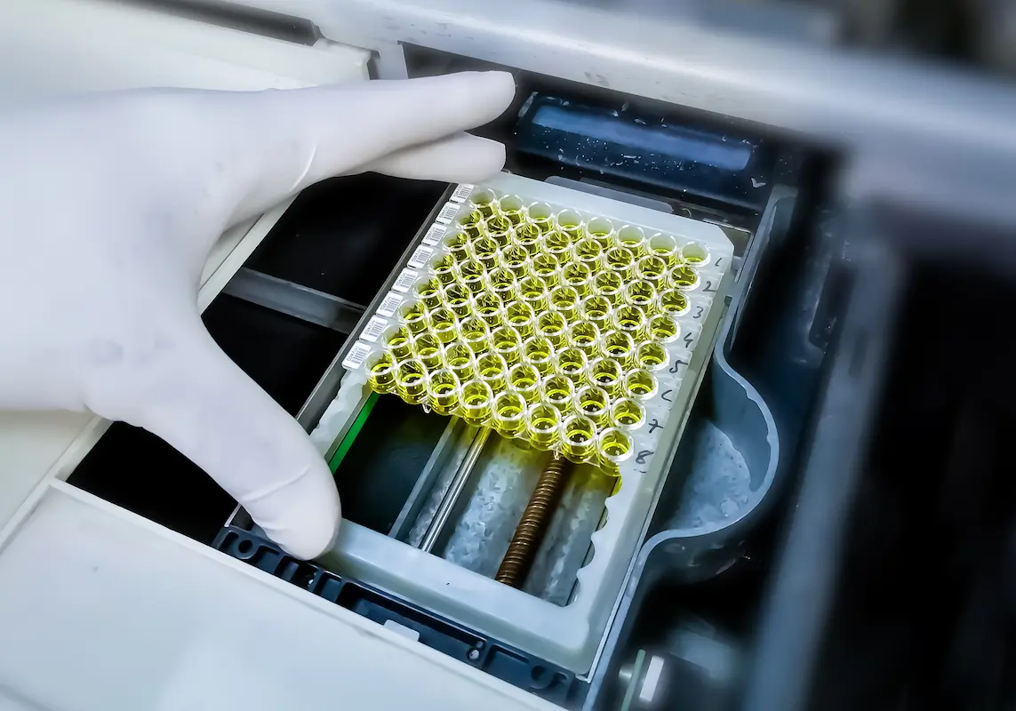A gloved hand is placing a tray filled with yellow liquid samples into a laboratory machine.