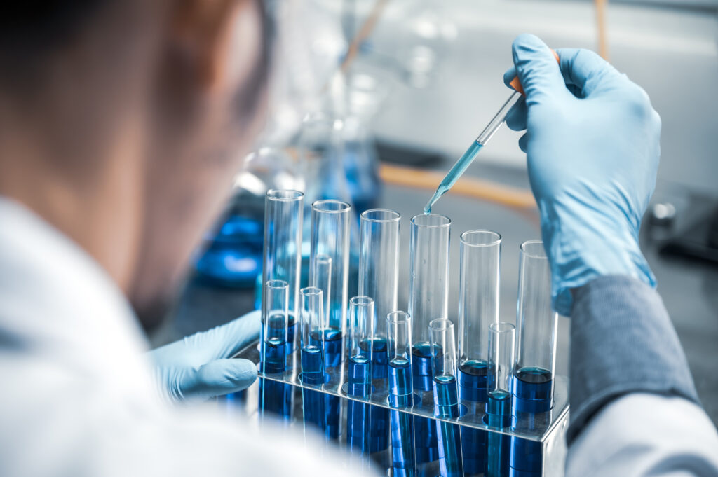 A scientist wearing gloves uses a pipette to add liquid to a row of test tubes filled with blue liquid in a laboratory setting.