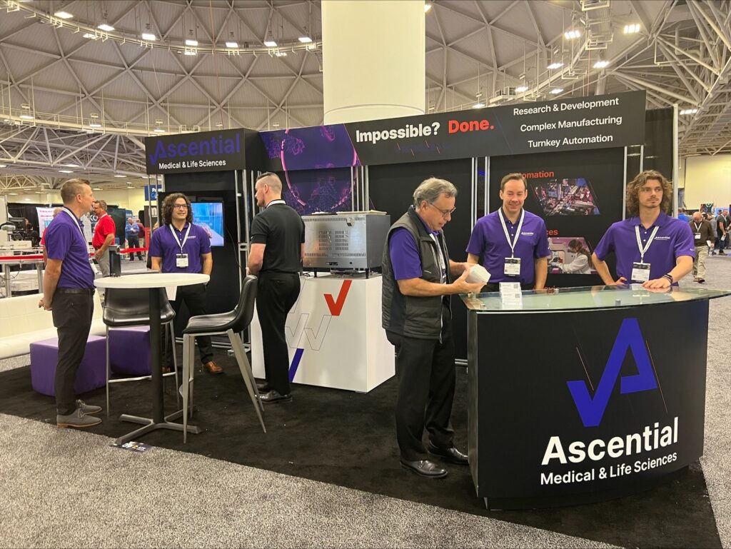 People in branded shirts interact at an Ascential Medical & Life Sciences trade show booth with displays and brochures.