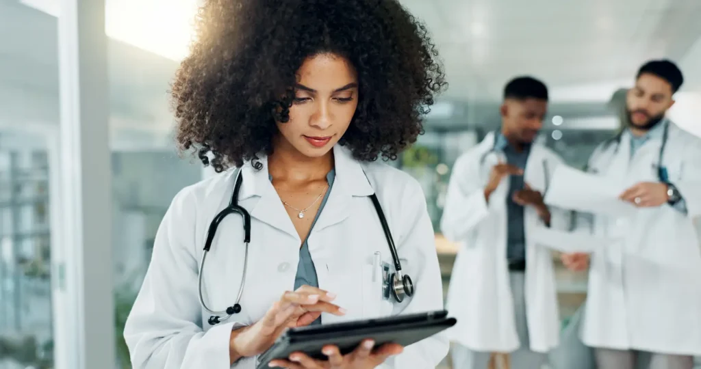 A doctor with a stethoscope uses a tablet in the foreground, while two other doctors review documents in the background of a medical office.